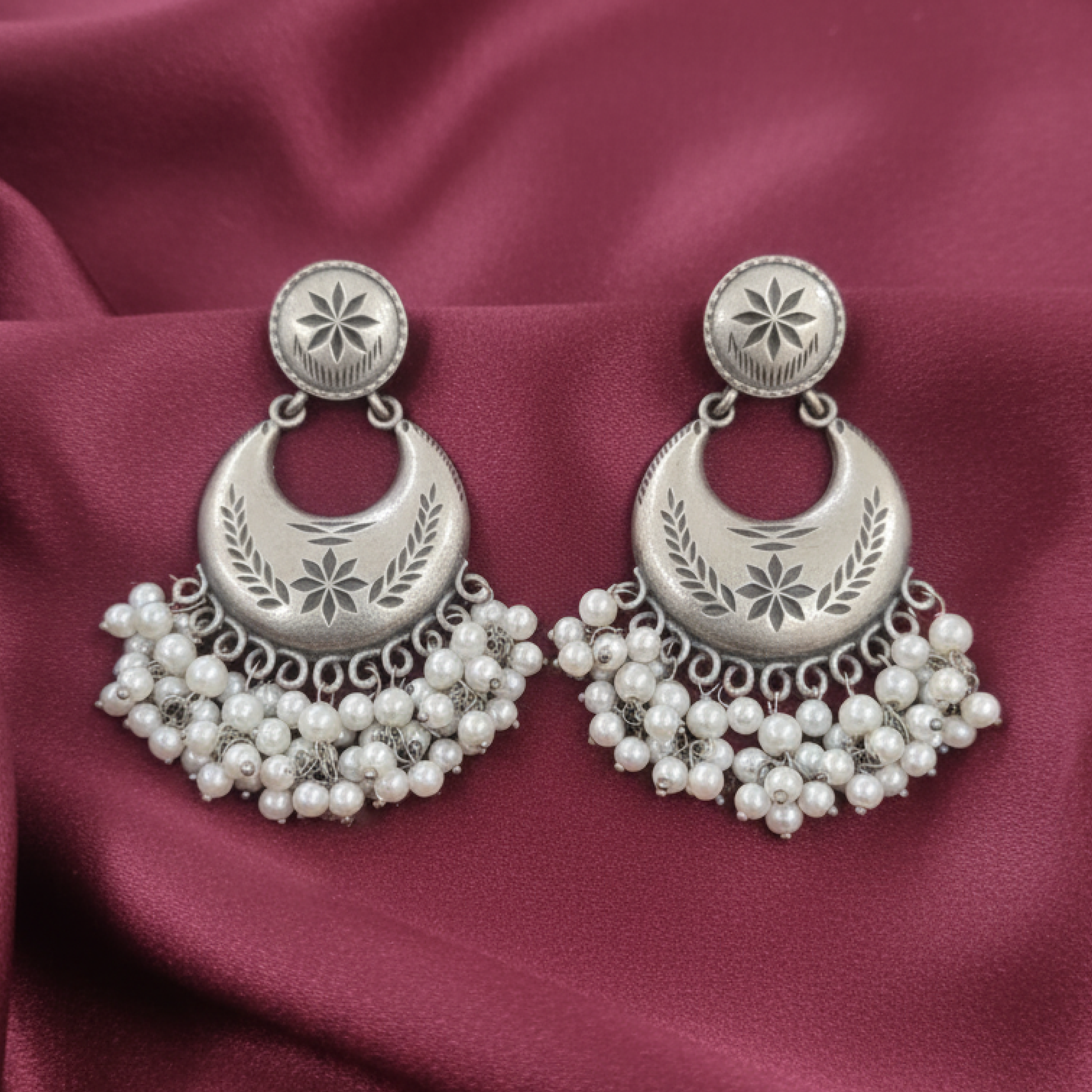 Oxidized silver crescent earrings with engraved detailing and pearl hangings displayed on a black stand white