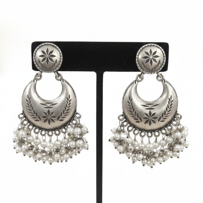 Oxidized silver crescent earrings with engraved detailing and pearl hangings displayed on a black stand ground