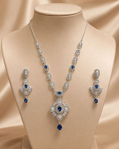 Elegant silver-plated necklace with large blue and clear stone pendant displayed on beige mannequin bust alongside matching blue teardrop earrings against soft beige fabric background 1