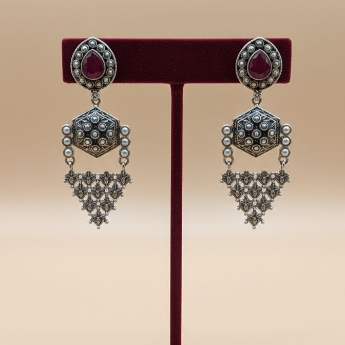 Pair of traditional Indian oxidized silver drop earrings with red teardrop gemstones, hexagonal middle motifs and intricate filigree triangular danglers on white background 3