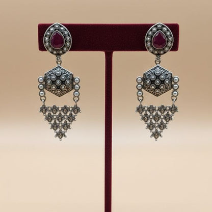 Pair of traditional Indian oxidized silver drop earrings with red teardrop gemstones, hexagonal middle motifs and intricate filigree triangular danglers on white background 3