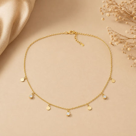 Delicate gold necklace with multiple circular charms and three clear crystal pendants displayed on beige fabric background 1