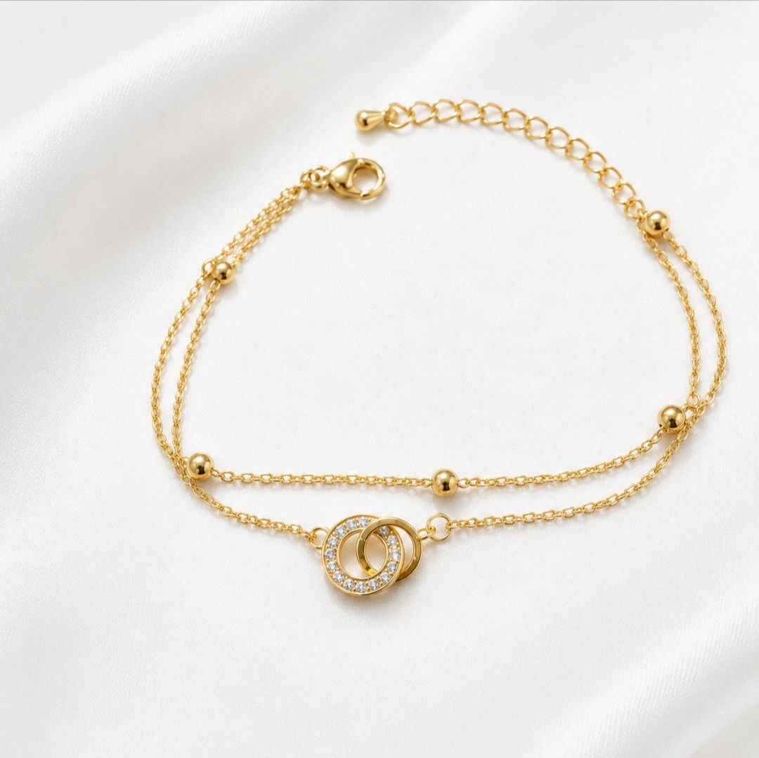 Premium gold bracelet with two interlocking circles one encrusted with sparkling crystals on delicate chain with gold bead accents displayed on white fabric 1