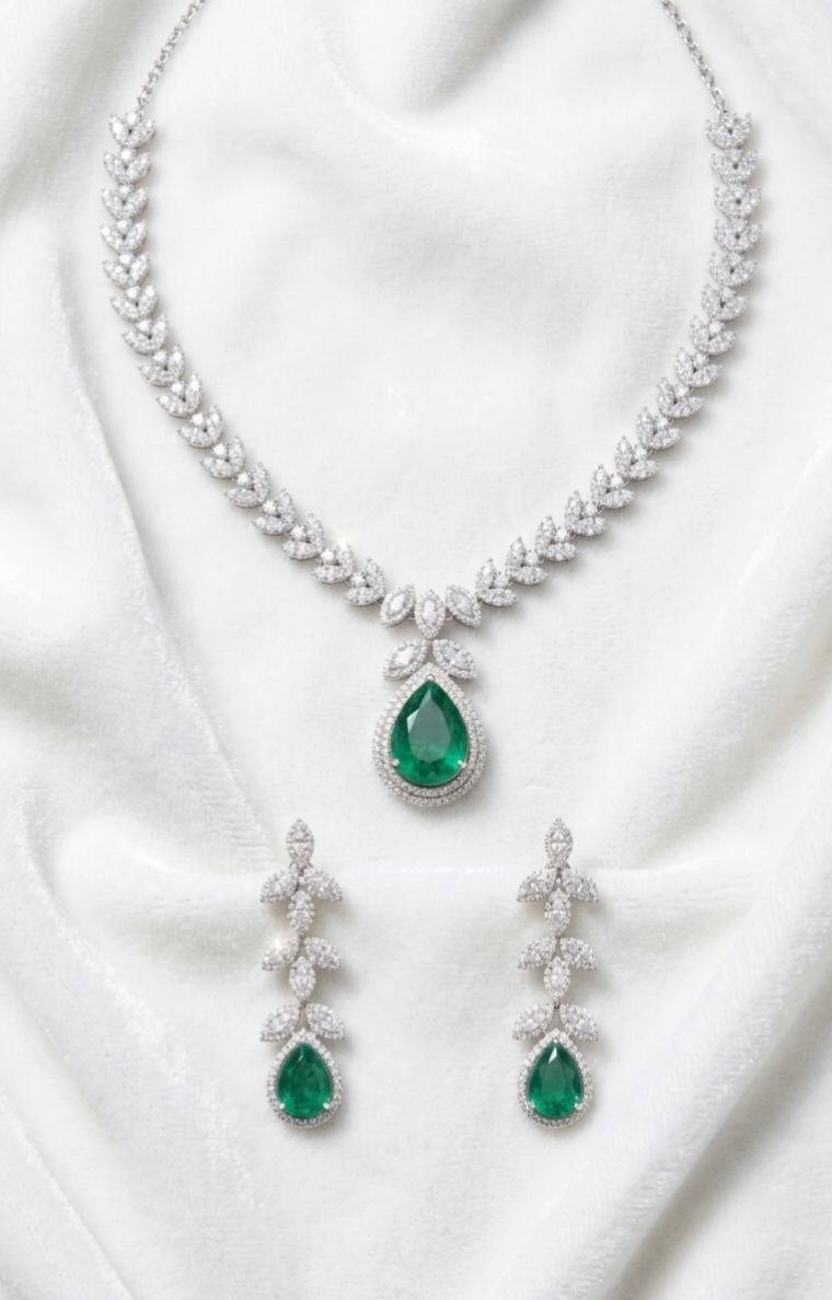 Luxury silver necklace with large green emerald teardrop pendant and matching dangling earrings featuring leaf motif cubic zirconia stones on white fabric 1