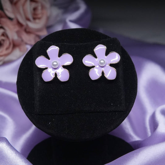 A pair of vibrant lavender enamel flower-shaped stud earrings, each with a small pearl in the center, displayed on a black velvet earring holder against a soft purple and pink silk background.
