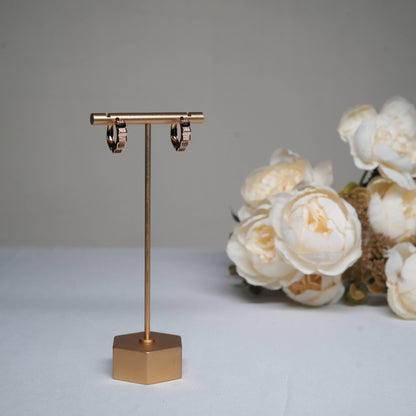 A pair of small, elegant rose gold-toned hoop earrings, embellished with sparkling pave diamonds, displayed on a minimalist gold-toned T-bar earring stand against a light grey background.