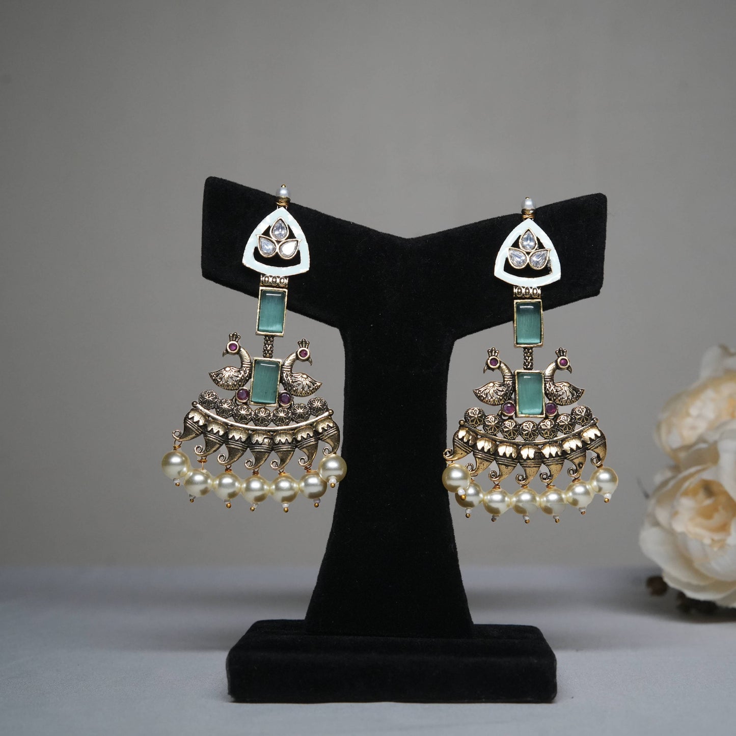 Antique gold temple earrings with kundan stones, emerald green accents, and pearl drops, featuring intricate peacock motifs displayed on a black velvet stand.