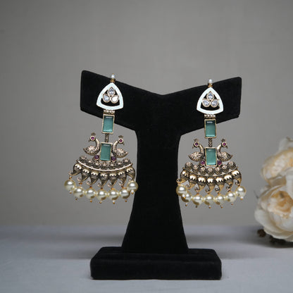 Antique gold temple earrings with kundan stones, emerald green accents, and pearl drops, featuring intricate peacock motifs displayed on a black velvet stand.