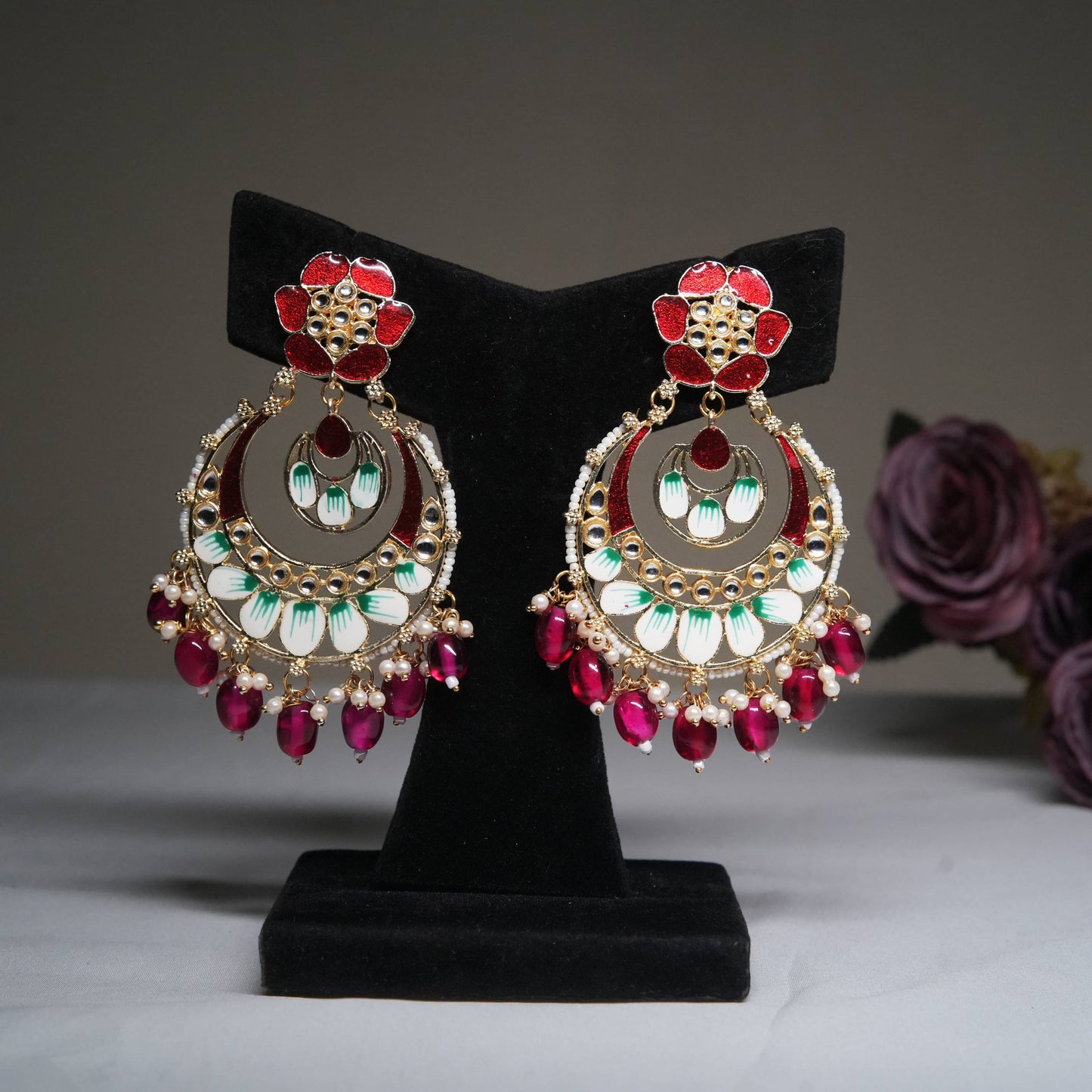 Enamel red and green chandbali earrings with ruby bead tassels displayed on a black velvet stand.