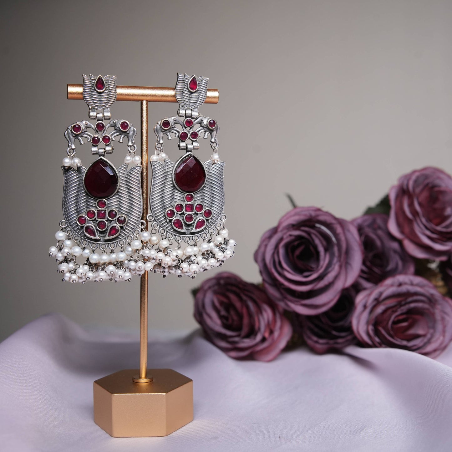 Ruby red temple earrings with oxidized silver finish and pearl tassels displayed on a golden stand.
