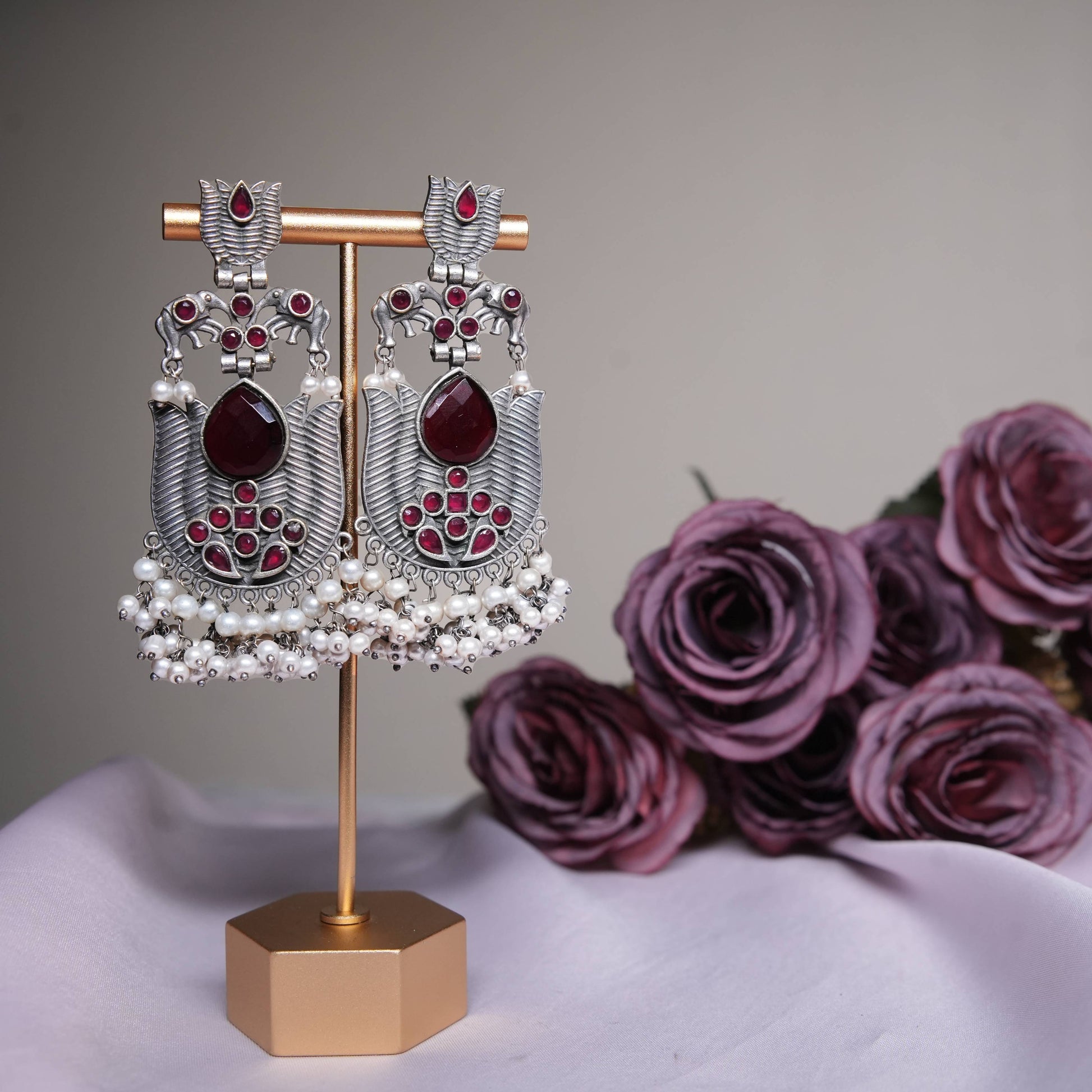 Ruby red temple earrings with oxidized silver finish and pearl tassels displayed on a golden stand.