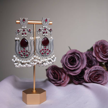 Ruby red temple earrings with oxidized silver finish and pearl tassels displayed on a golden stand.